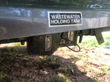 RV rental's wastewater holding tank with a Valterra control component and attached power cord, seen from below near the ground.