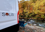 Solis RV rental by an autumn river. A camping chair offers views of colorful trees and flowing water.