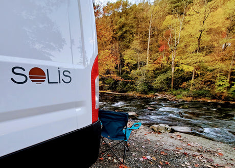 Solis RV rental by an autumn river. A camping chair offers views of colorful trees and flowing water.