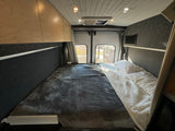 Cozy RV rental interior with a bed, overhead wood cabinets, and rear windows. Experience comfort in this camper van.