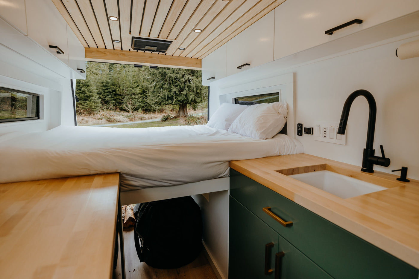 This RV rental features a cozy bed looking out to a serene forest view, with a compact kitchen and wood counters. — photo 13