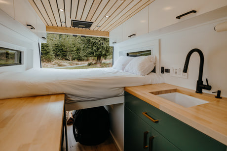 This RV rental features a cozy bed looking out to a serene forest view, with a compact kitchen and wood counters.