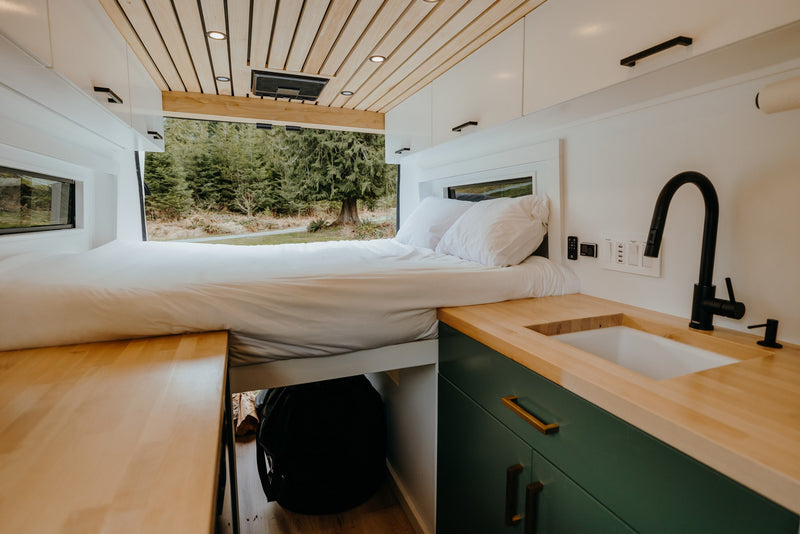 This RV rental features a cozy bed looking out to a serene forest view, with a compact kitchen and wood counters. — photo 13