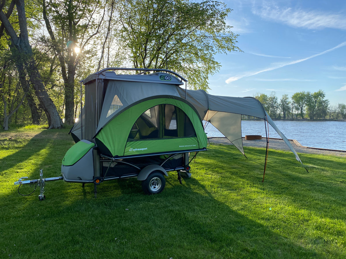 This compact SylvanSport trailer camper, ideal for an RV rental, is set up on lush green grass beside a scenic lake.