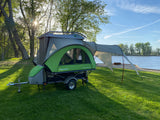 This compact SylvanSport trailer camper, ideal for an RV rental, is set up on lush green grass beside a scenic lake.