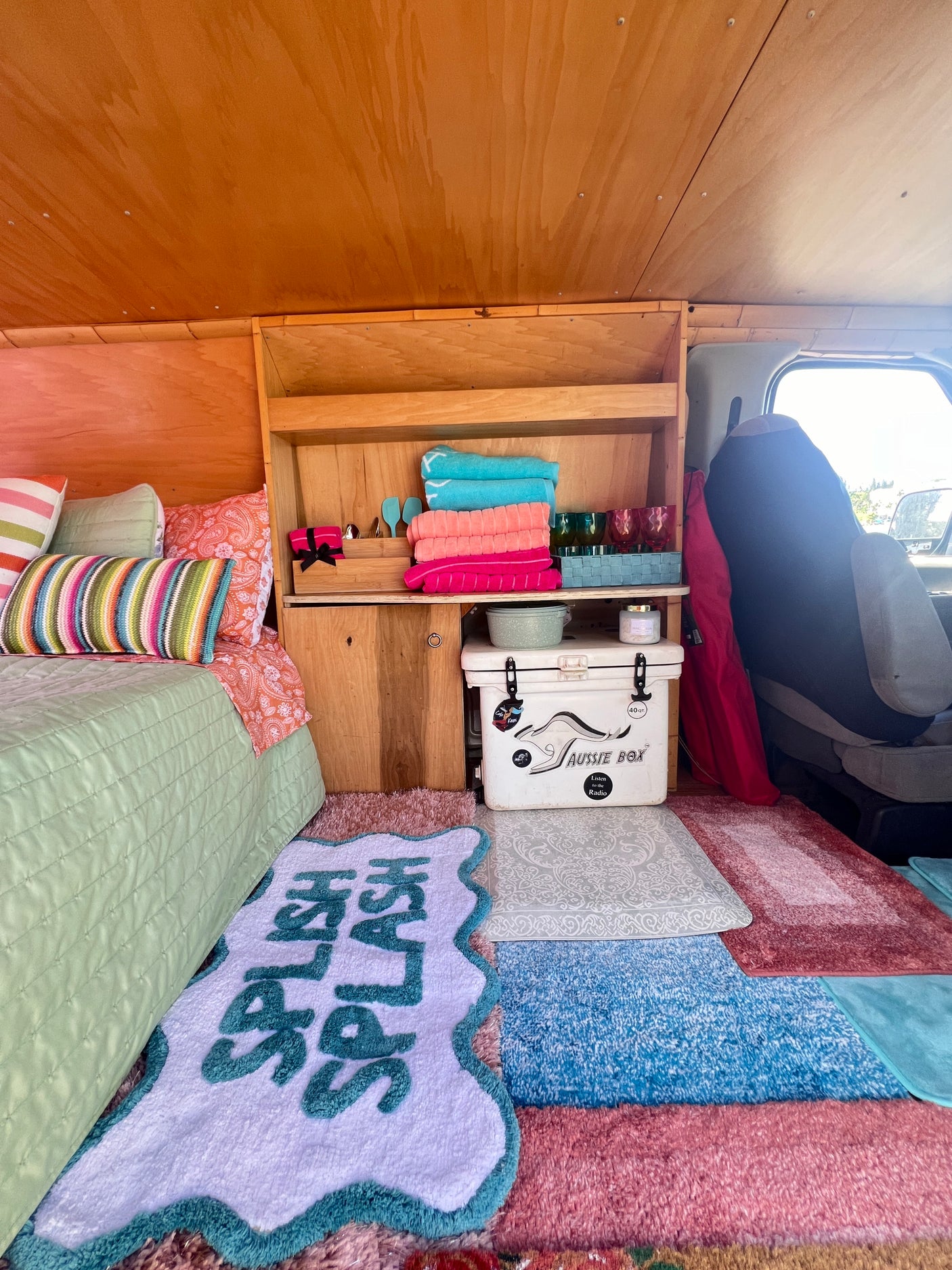 Vibrant RV rental interior: colorful bed, custom wood shelving with towels & cups, cooler, and playful rugs.