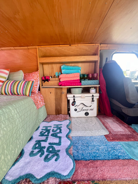Vibrant RV rental interior: colorful bed, custom wood shelving with towels & cups, cooler, and playful rugs.