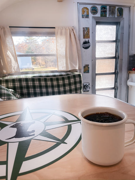 Coffee in an RV rental, showing a cozy interior with plaid seating, a compass table, and a window view of autumn trees.
