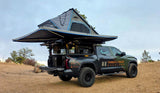 Dark green overland truck with deployed rooftop tent & awning on a dirt path. Ideal RV rental for off-grid camping adventures.