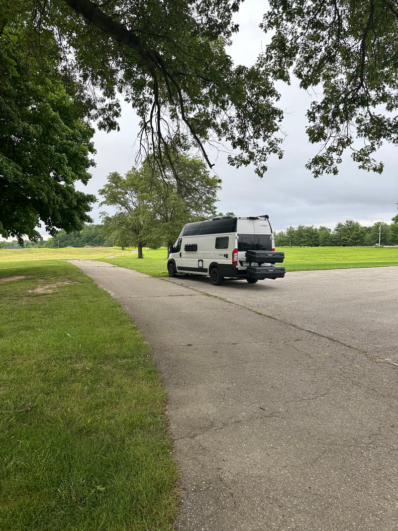 An RV rental - a white Noovo camper van with storage boxes - parked on a paved path next to grass and trees under a cloudy sky. — photo 11