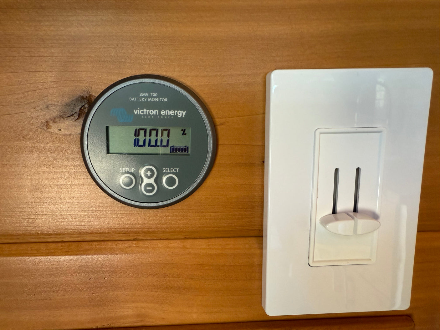 RV rental's Victron Energy battery monitor shows 100% charge next to a light switch on wooden paneling.