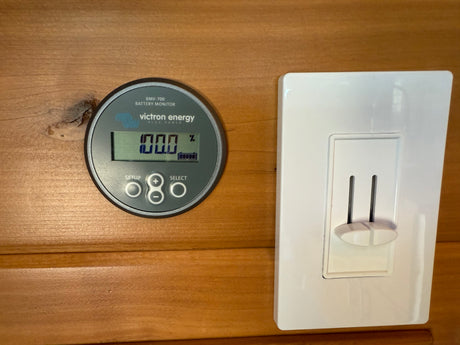 RV rental's Victron Energy battery monitor shows 100% charge next to a light switch on wooden paneling.