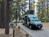 RV rental parked at a shaded forest campsite. A person works on a bike near the pop-top camper van, ready for outdoor adventures.