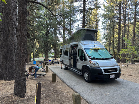 RV rental parked at a shaded forest campsite. A person works on a bike near the pop-top camper van, ready for outdoor adventures.