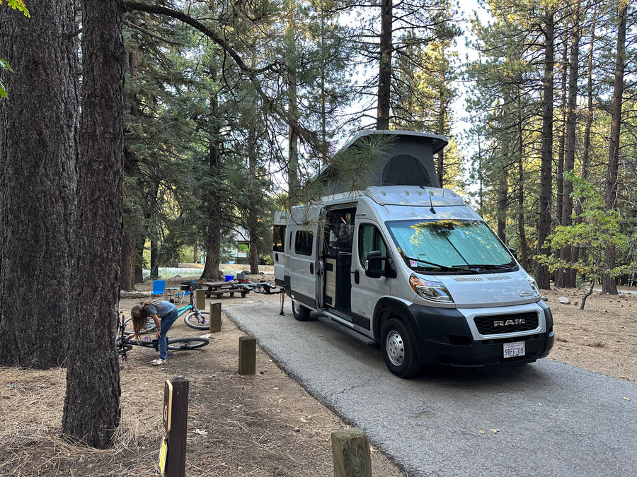 RV rental parked at a shaded forest campsite. A person works on a bike near the pop-top camper van, ready for outdoor adventures.
