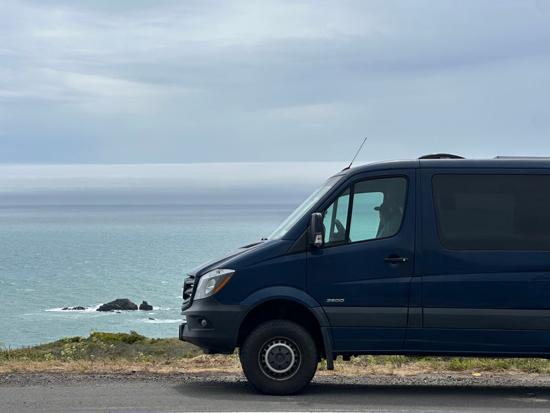  parked on a scenic California coast road overlooking cliffs & ocean under a cloudy sky.|Dark blue Mercedes Sprinter rv rental parked by a coastal road — photo 2