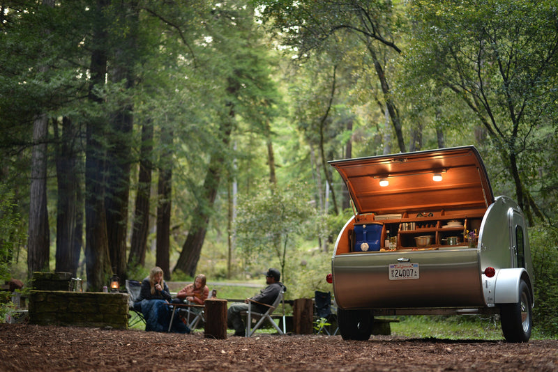 Experience camping comfort! This RV rental teardrop camper's rear kitchenette is open at a lush forest campsite with people relaxing. — photo 11