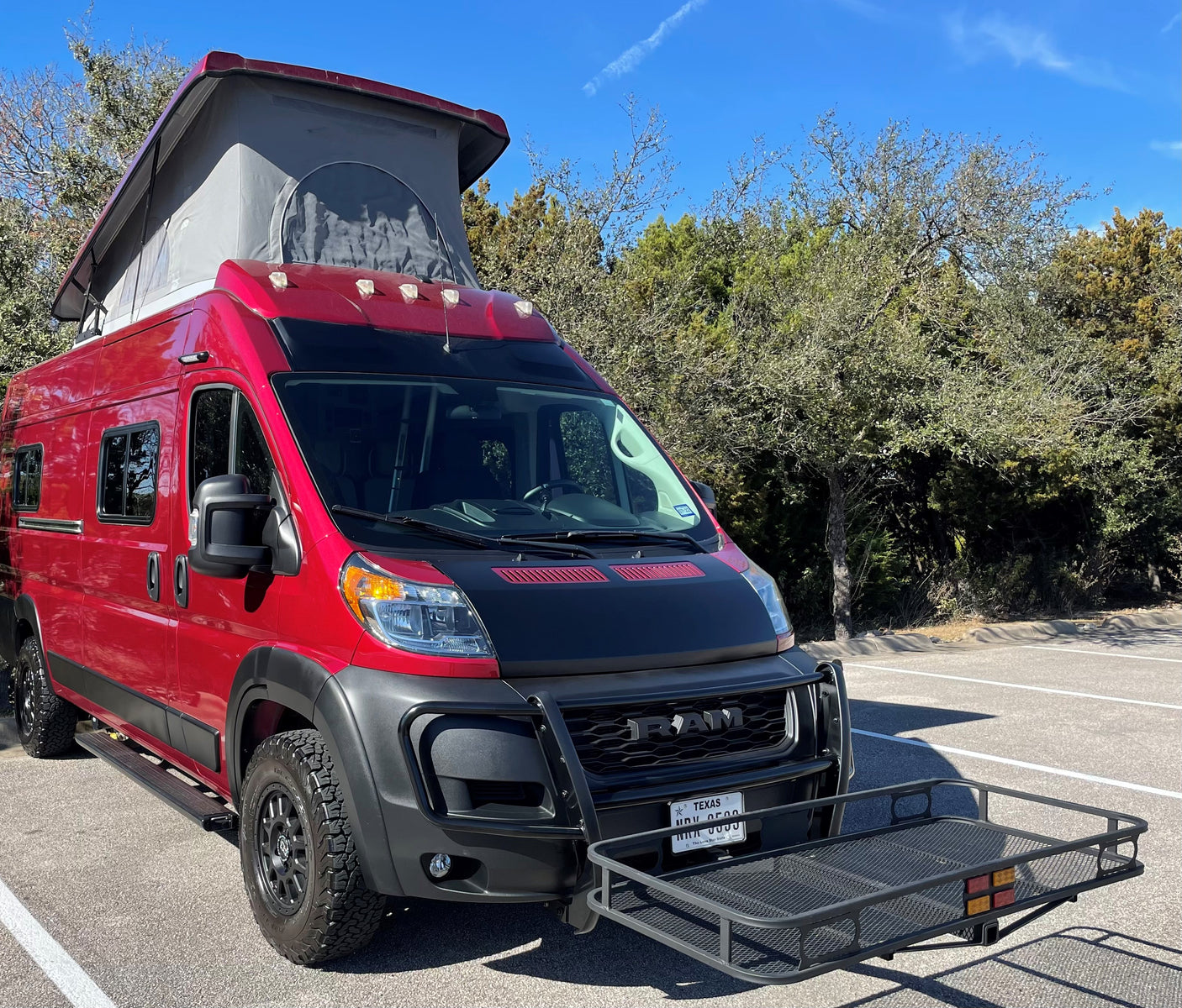 Red pop-top RV rental with off-road features, front cargo carrier, and Texas plates, parked outdoors.