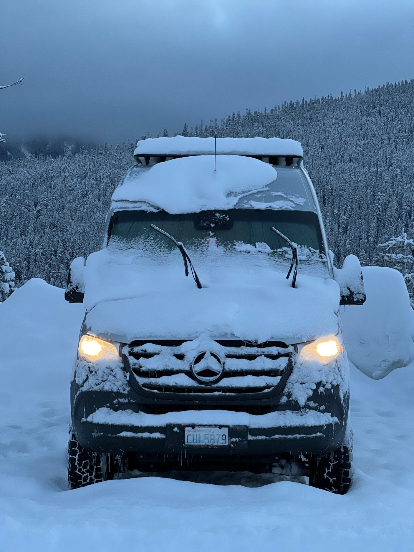 This Mercedes Sprinter RV rental, covered in snow, lights up a winter forest, ready for cold-weather adventures.