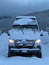 This Mercedes Sprinter RV rental, covered in snow, lights up a winter forest, ready for cold-weather adventures.