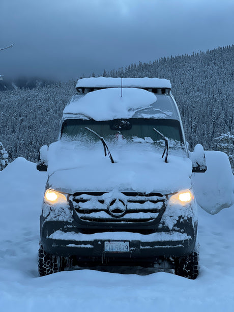 This Mercedes Sprinter RV rental, covered in snow, lights up a winter forest, ready for cold-weather adventures.