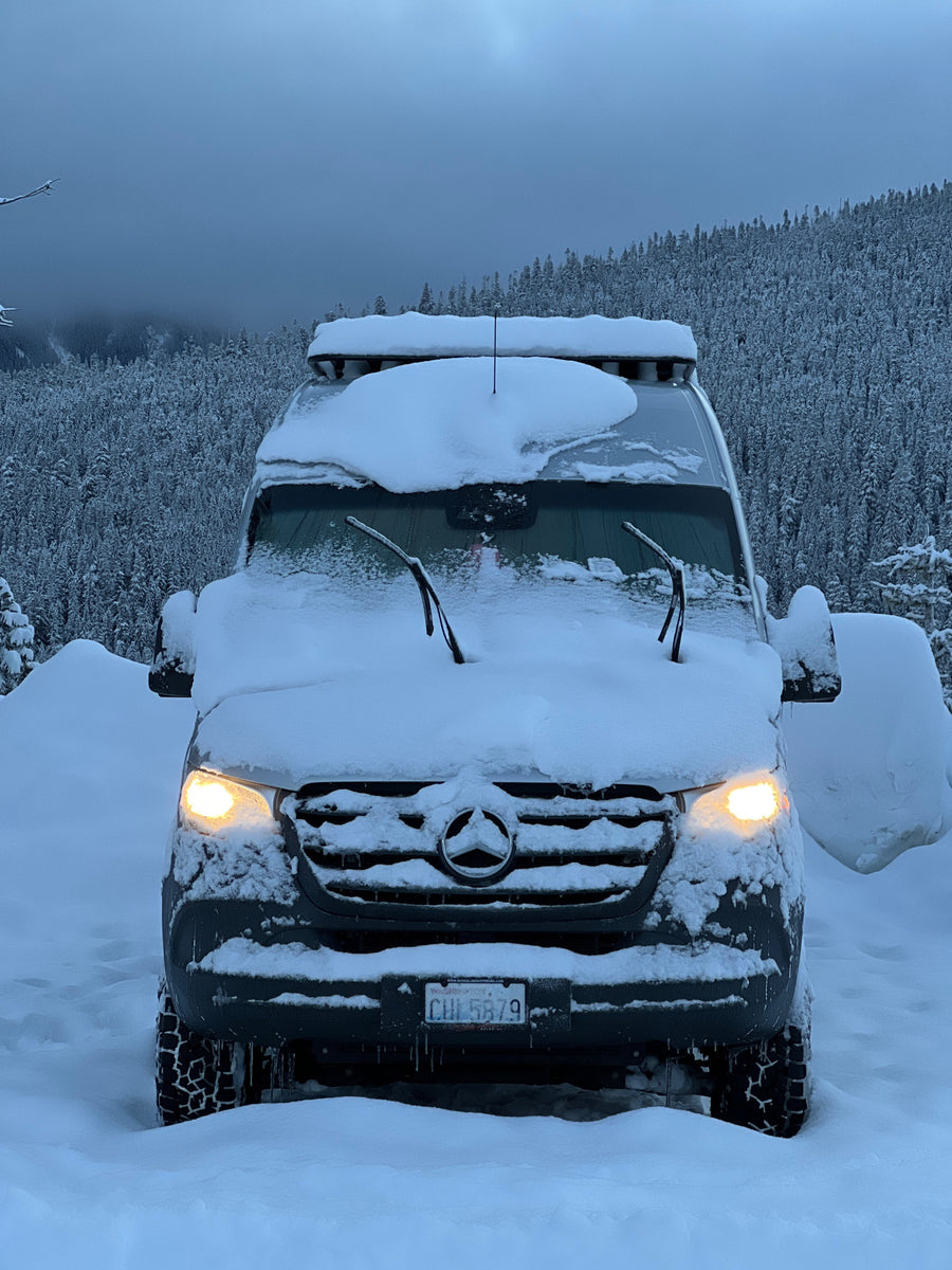 This Mercedes Sprinter RV rental, covered in snow, lights up a winter forest, ready for cold-weather adventures.