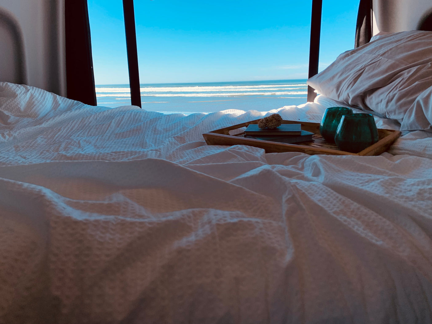 Cozy RV rental bedroom offers stunning ocean views! White bed with a tray, books, and glasses for a serene escape.