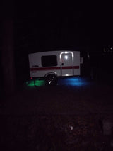 Compact teardrop RV rental at night, lit by vibrant green and blue ground effects, perfect for camping.