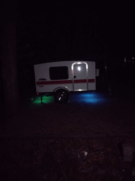 Compact teardrop RV rental at night, lit by vibrant green and blue ground effects, perfect for camping.