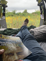 Enjoying an RV rental view of goldenrod fields while reading 'Outside' magazine's bucket list.