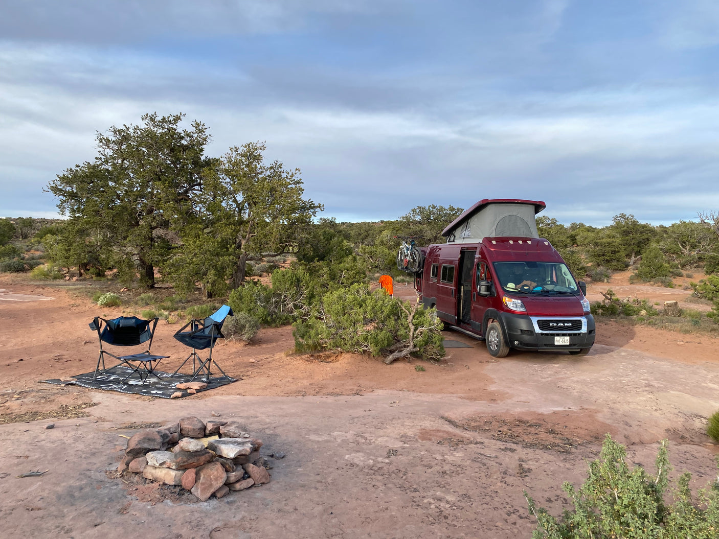 Red RV rental camper van parked in a spacious desert campsite with chairs and a fire pit under a cloudy sky.