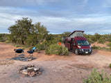 Red RV rental camper van parked in a spacious desert campsite with chairs and a fire pit under a cloudy sky.