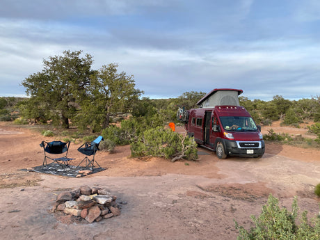 Red RV rental camper van parked in a spacious desert campsite with chairs and a fire pit under a cloudy sky.