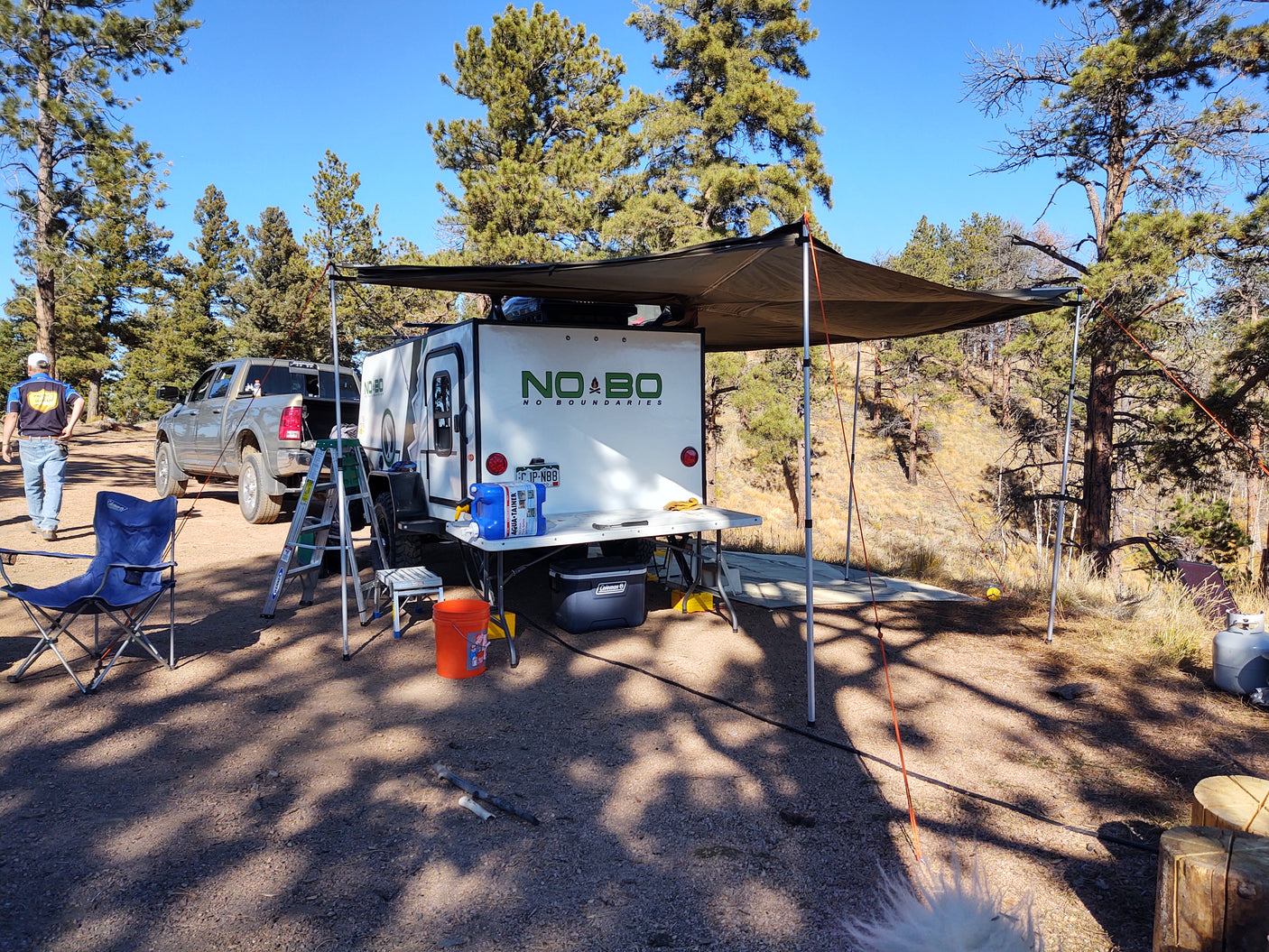 White NO BO rv rental camper set up at a sunny campsite with an awning, truck, and pine trees.