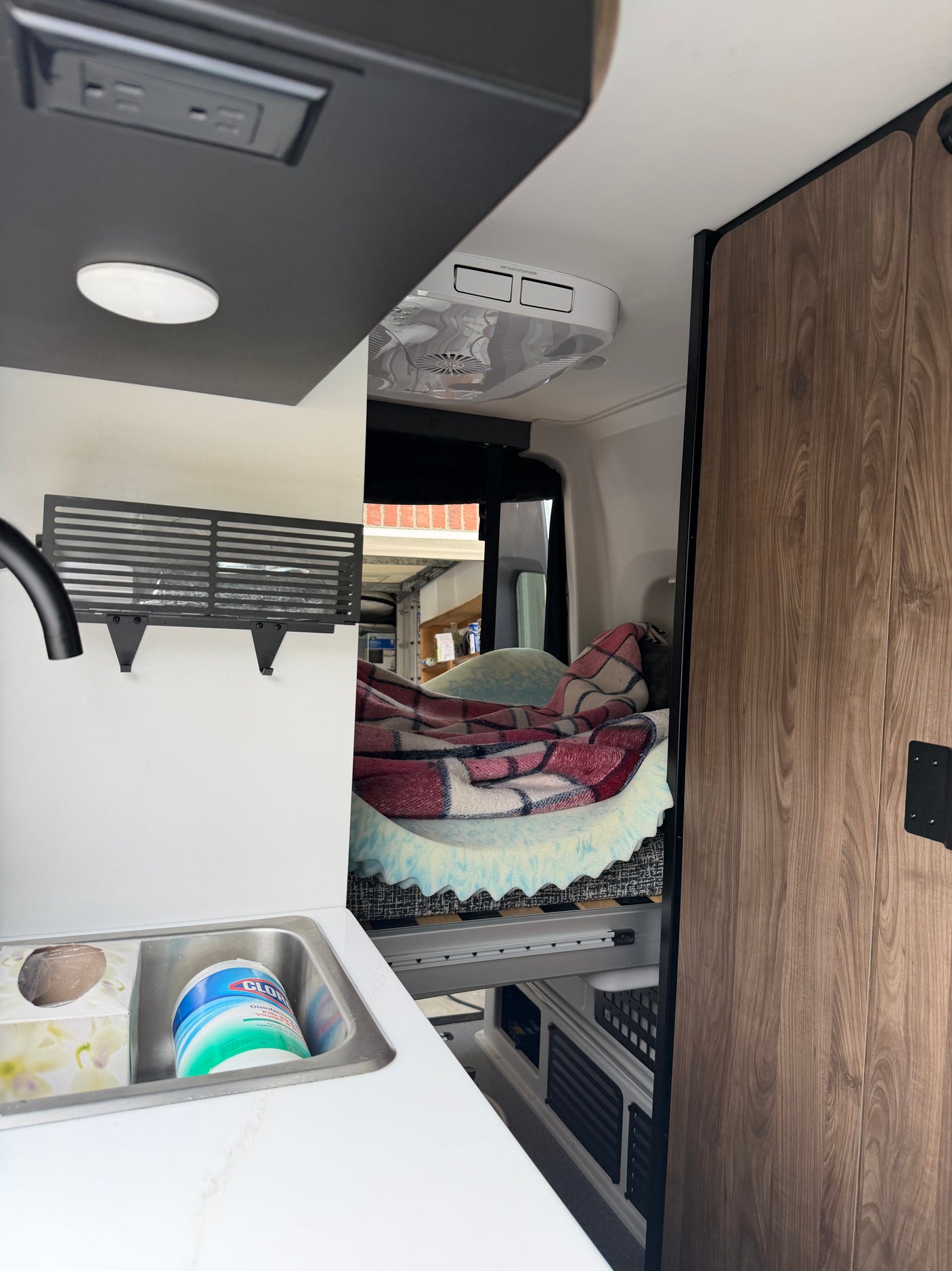 Practical rv rental interior: view of a compact sink with cleaning supplies, overhead power, and a cozy bed area.