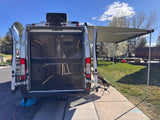 Spacious RV rental, seen from the rear with open doors, mesh screen, and extended awning over chairs. Interior bed visible for travel.