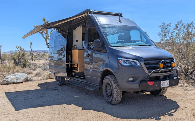 Dark grey camper van with extended awning in a sunny desert landscape with Joshua trees. Great for an rv rental adventure!