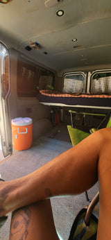 RV rental interior features a bed, orange cooler, and camping chairs, viewed from a person's seated perspective.