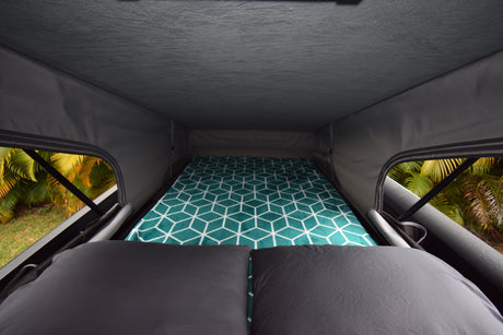 Inside an RV rental pop-top: a comfortable bed with teal patterned blanket & dark pillows, overlooking green scenery.