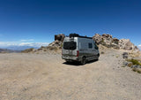 Silver Winnebago Revel camper van, possibly an RV rental, parked on a rocky desert overlook with distant mountains.
