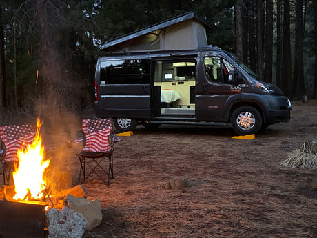 Enjoying a cozy evening camping with an RV rental. A dark grey pop-top camper van, American flag chairs, and a bright campfire at dusk.