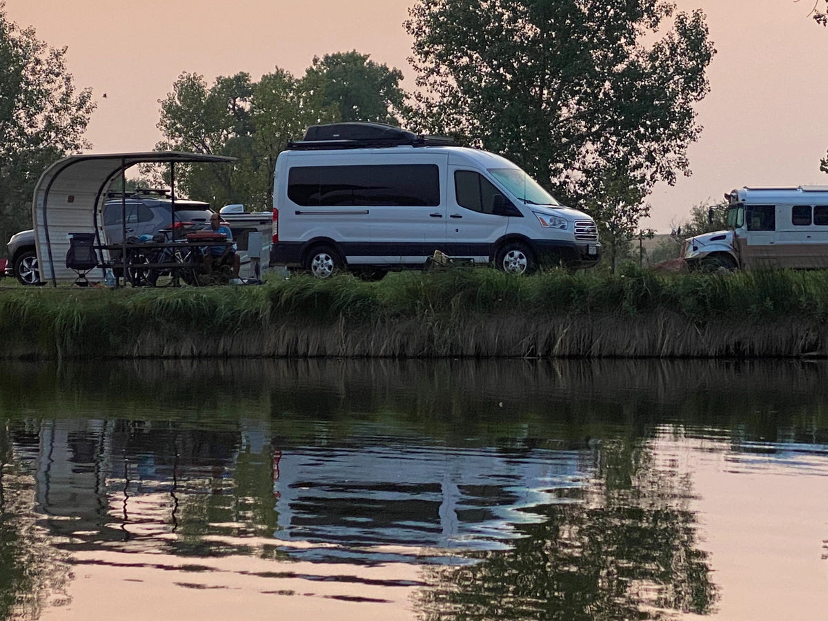 Evening view of a riverside campsite featuring an RV rental van, SUV, and RV with reflections in the water.