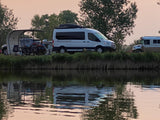 Evening view of a riverside campsite featuring an RV rental van, SUV, and RV with reflections in the water.