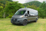A dark grey customized camper van, suitable for rv rental, parked on a green grassy field with lush trees & hills behind.