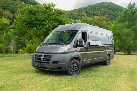 A dark grey customized camper van, suitable for rv rental, parked on a green grassy field with lush trees & hills behind.