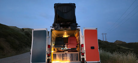 RV rental campervan's rear open, revealing two chairs, warm lights & rooftop tent, set against dusk hills.