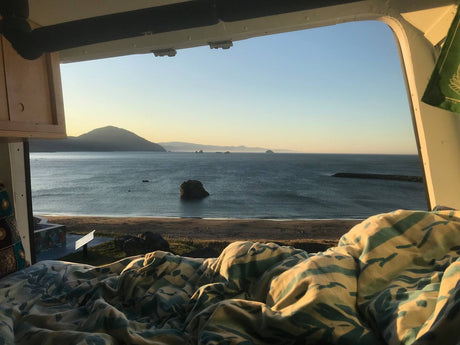Cozy RV rental bed with a stunning ocean view at sunrise, overlooking a bay, distant mountains, and a large sea stack.