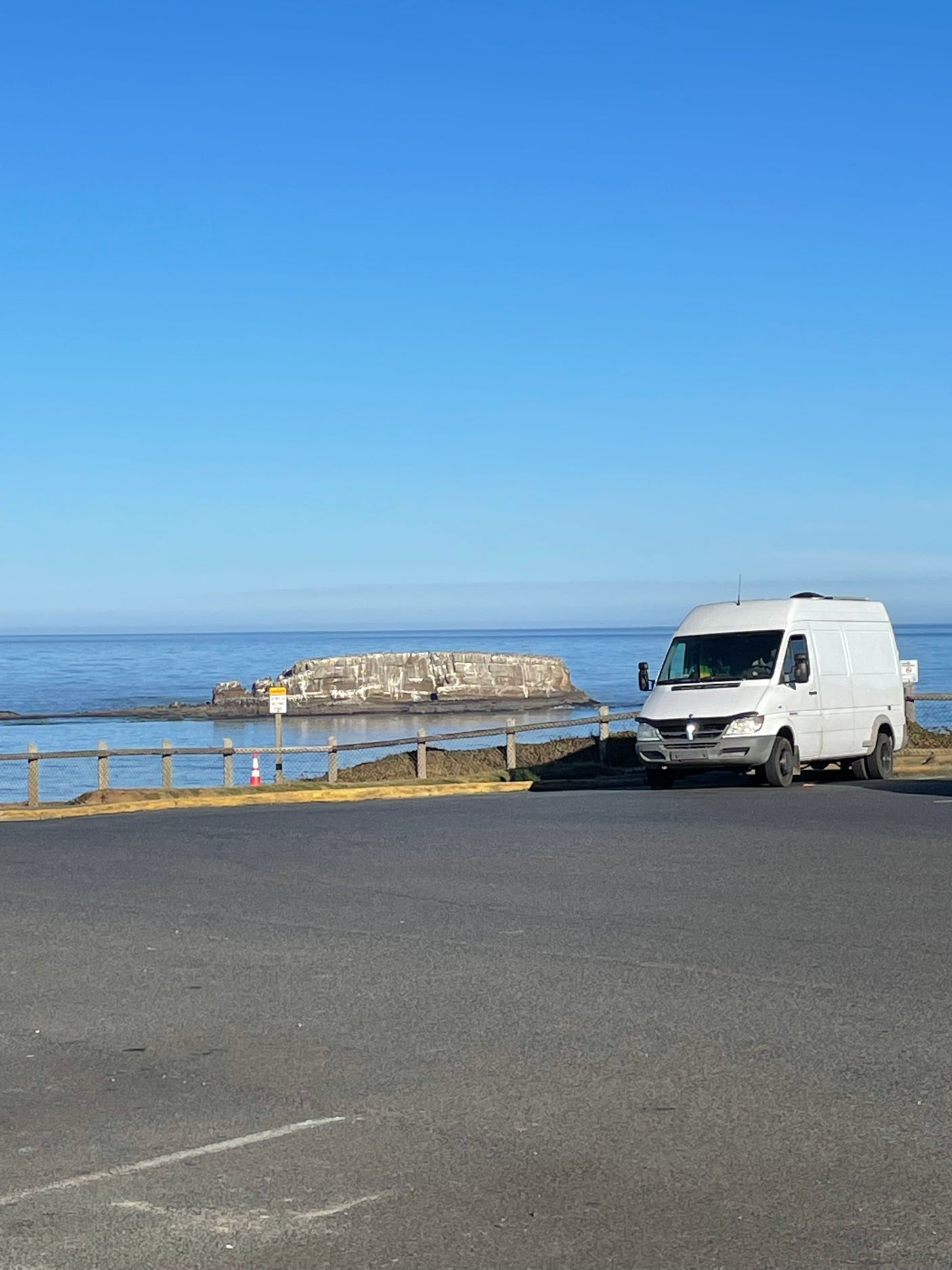 White RV rental van parked on a coastal road overlooking the ocean & cliffs under a clear blue sky.