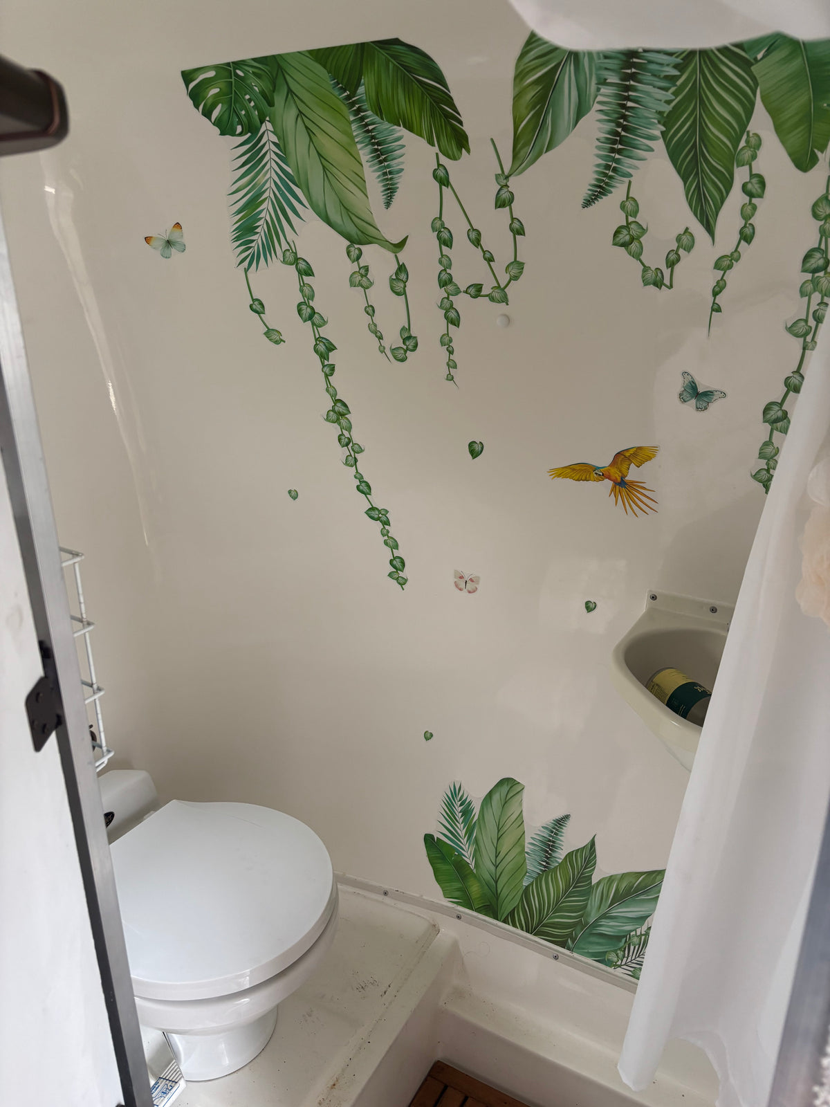 RV rental bathroom with tropical leaf, butterfly, and parrot decals. Features a white toilet, compact sink, and shower area. — photo 7