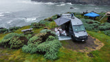 RV rental van parked on a coastal cliff, awning extended. People enjoy the ocean view in this scenic outdoor camp setup.